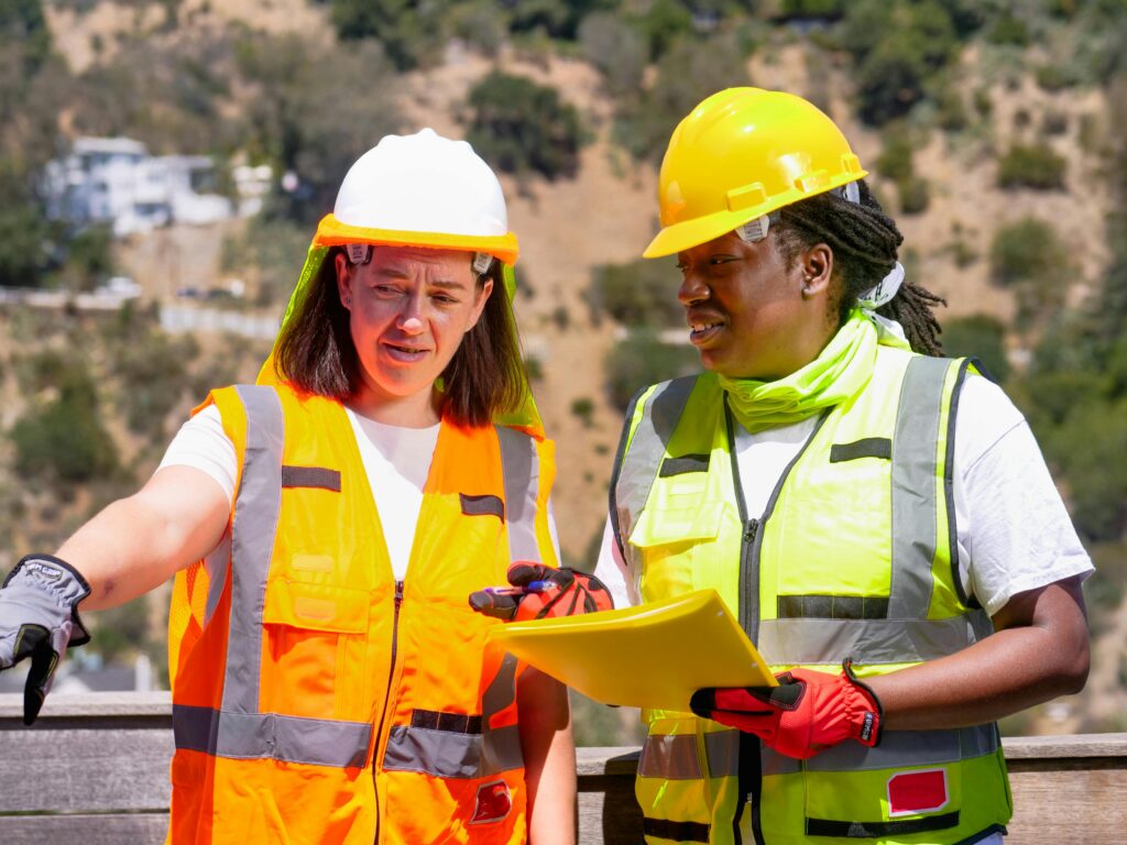 Two female engineers in safety gear discuss a project outdoors, focusing on plans.